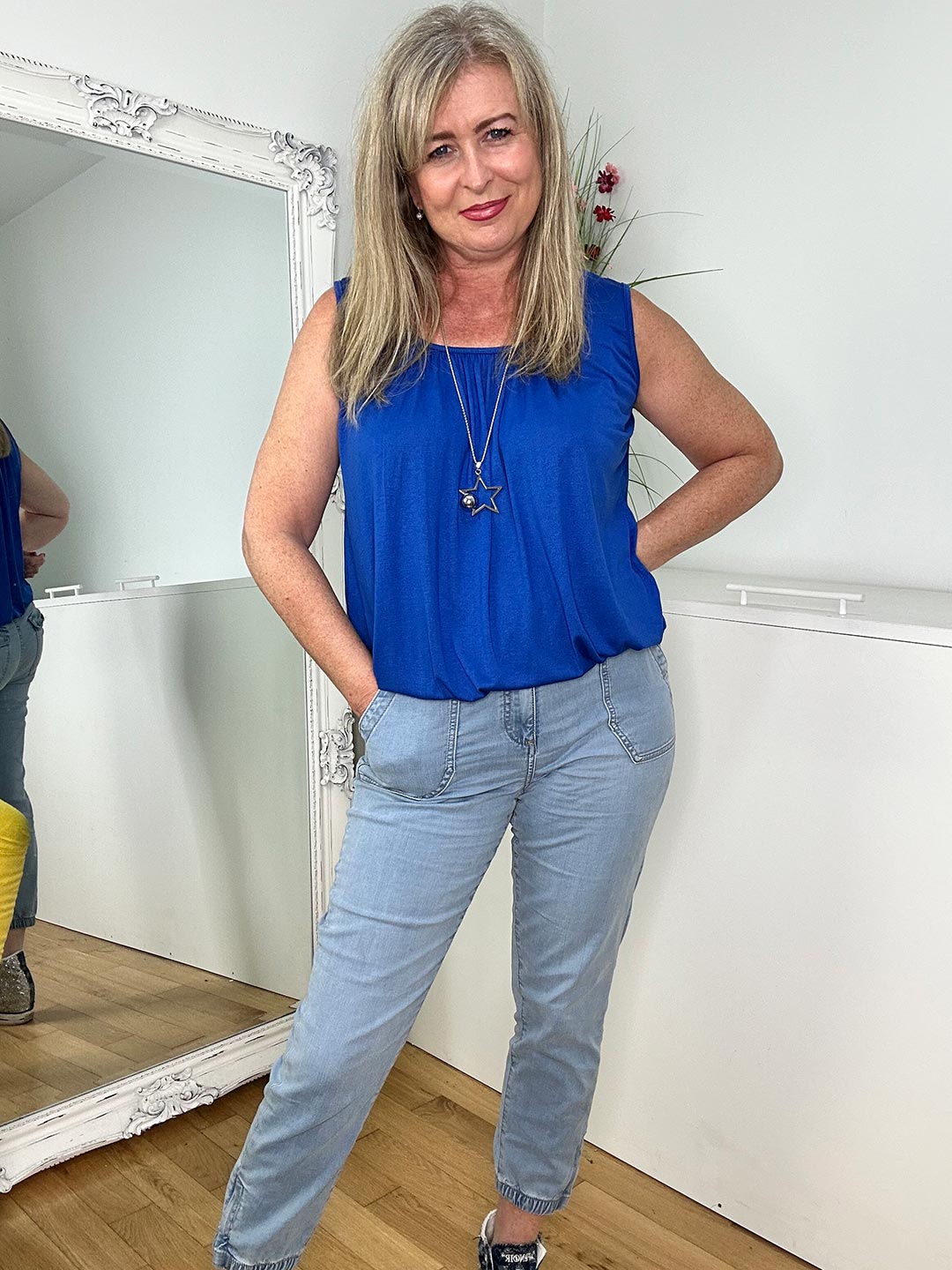 Woman wearing a blue sleeveless top and light blue jeans standing in front of a mirror.